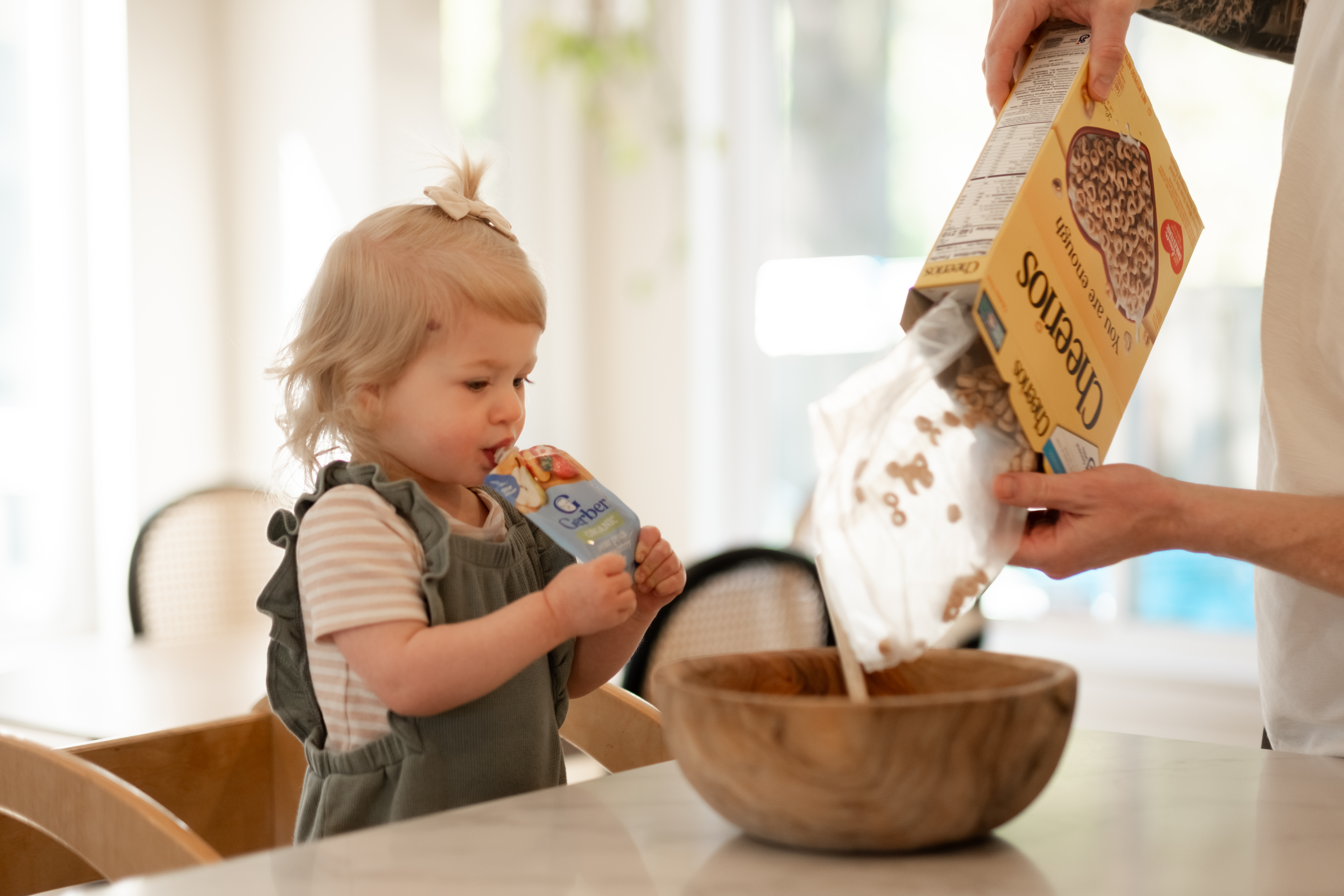 Picture of toddler girl standing on stool at the kitchen counter while eating a squeeze pack of Gerber puree